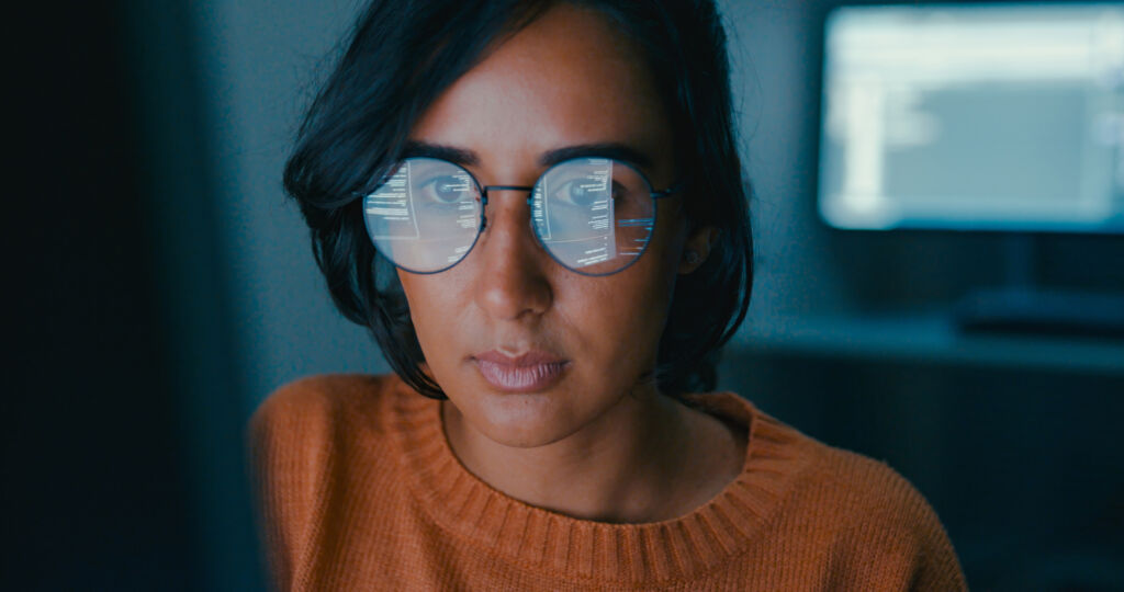 Female IT professional looking at screen in dark room with screen mirrored on her glasses